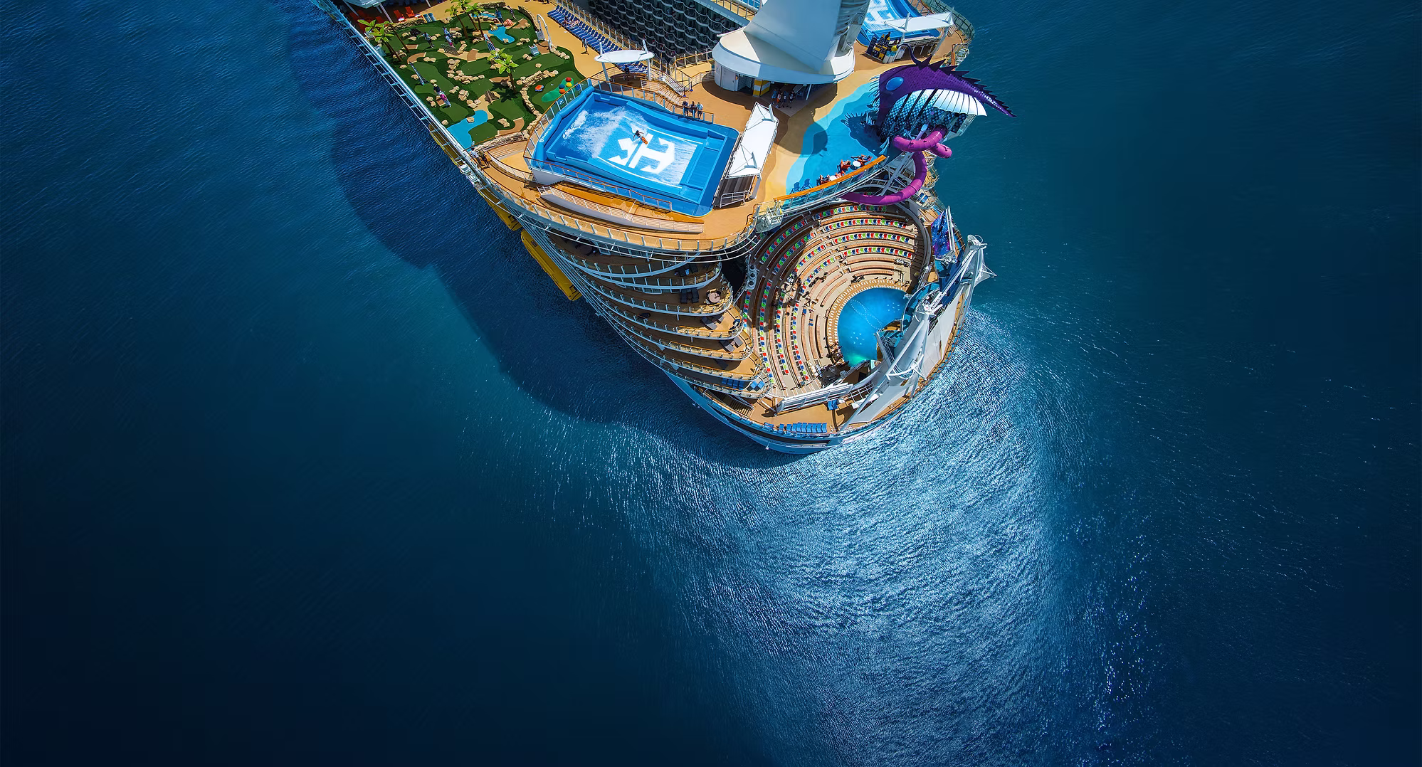 Aerial view of a large cruise ship's stern under way in open water, its top-deck pools and sports decks visible